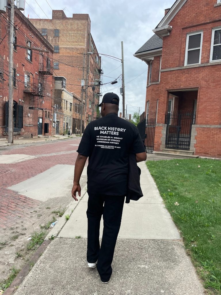 Image: Jamon Jordan, the first official City Historian of the City of Detroit, walks away from the camera. He is dressed in a black t-shirt with the words "Black History Matters" on the back, and black pants. He is walking down a sidewalk in Detroit. Photograph by Irina Zadov.