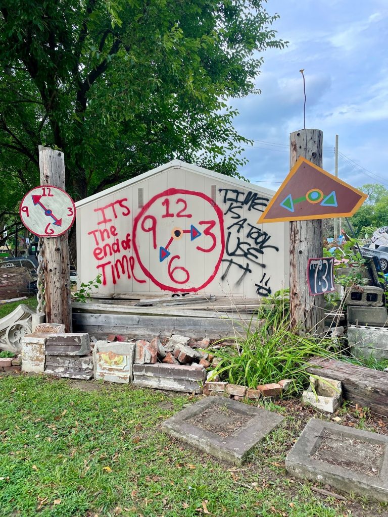 Image: A close up of the Tyree Guyton’s Heidelberg Project featuring a wooden structure in the shape of a small home or a large headstone. The wooden structure is painted white and on top of the paint there is a clock spray painted in black, red, and blue. The clock's hands point to the 3 and in between the 6 and 9. Around the clock, the spray paint reads: IT'S THE END OF TIME. TIME IS A TOOL, USE IT. Photograph by Irina Zadov.