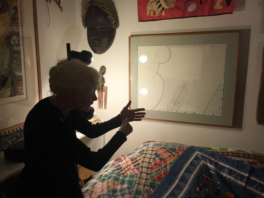 Chicago artist Barbara Rossi gestures to one of her works installed in the Roger Brown Study Collection. The artist is in shadow while a bright spotlight shines on the piece she's gesturing toward. The artwork is hung on a white wall surrounded by a member of other art pieces.