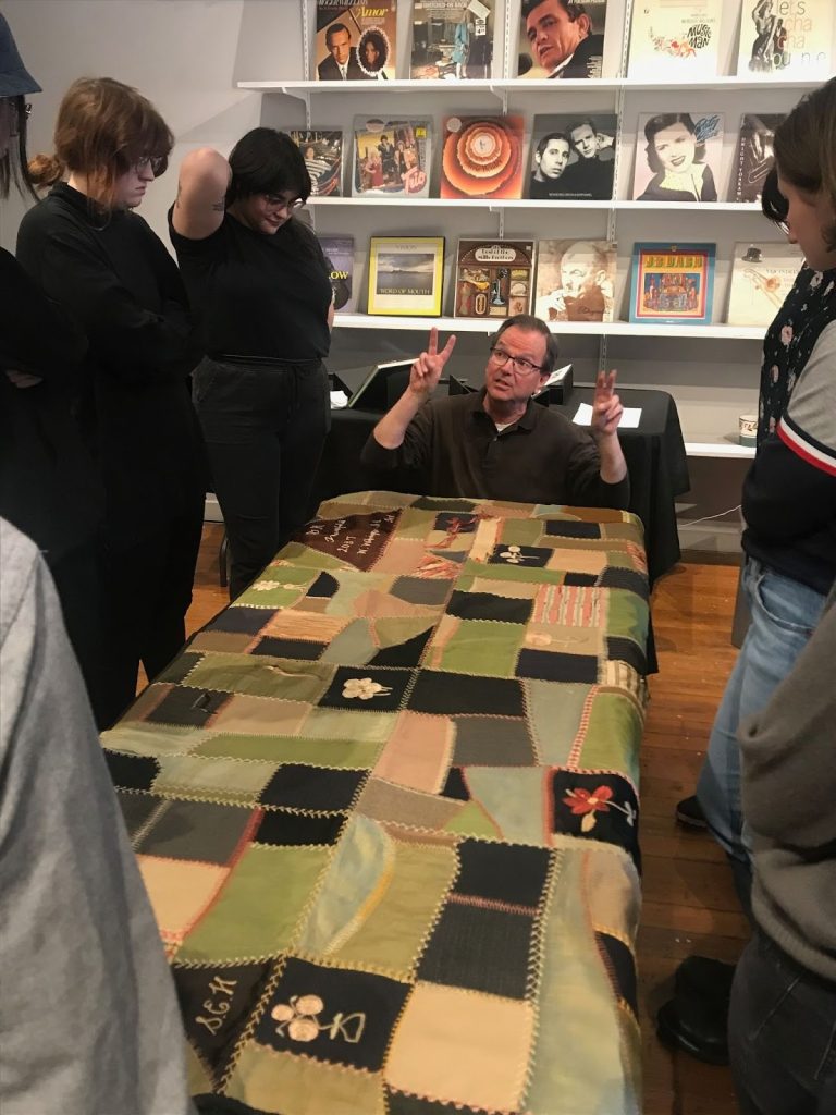 SAIC professor Jerry Bleem sits/kneels at the head of a  table upon which sits a large multicolored quilt. He's speaking to a group of people who look both at thim and at the quilt on display.
