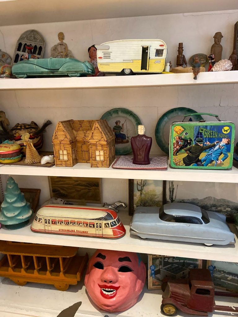 Four white inset shelves laden with various knickknacks and art works. On the bottom shelf is a pink mask sandwiched between a small truck and a trolly. Theres are candle shaped like a Christmas tree, a green later lunchbox and a miniature yellow airstream trailer. 