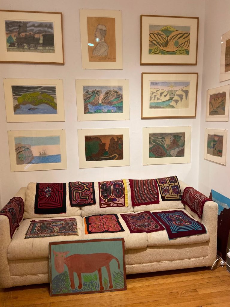 Joseph Yoakum room at the Roger Brown Study Collection. Nine prints hang on a white wall above a white couch draped in multicolored squares of quilted and embroidered fabric. 