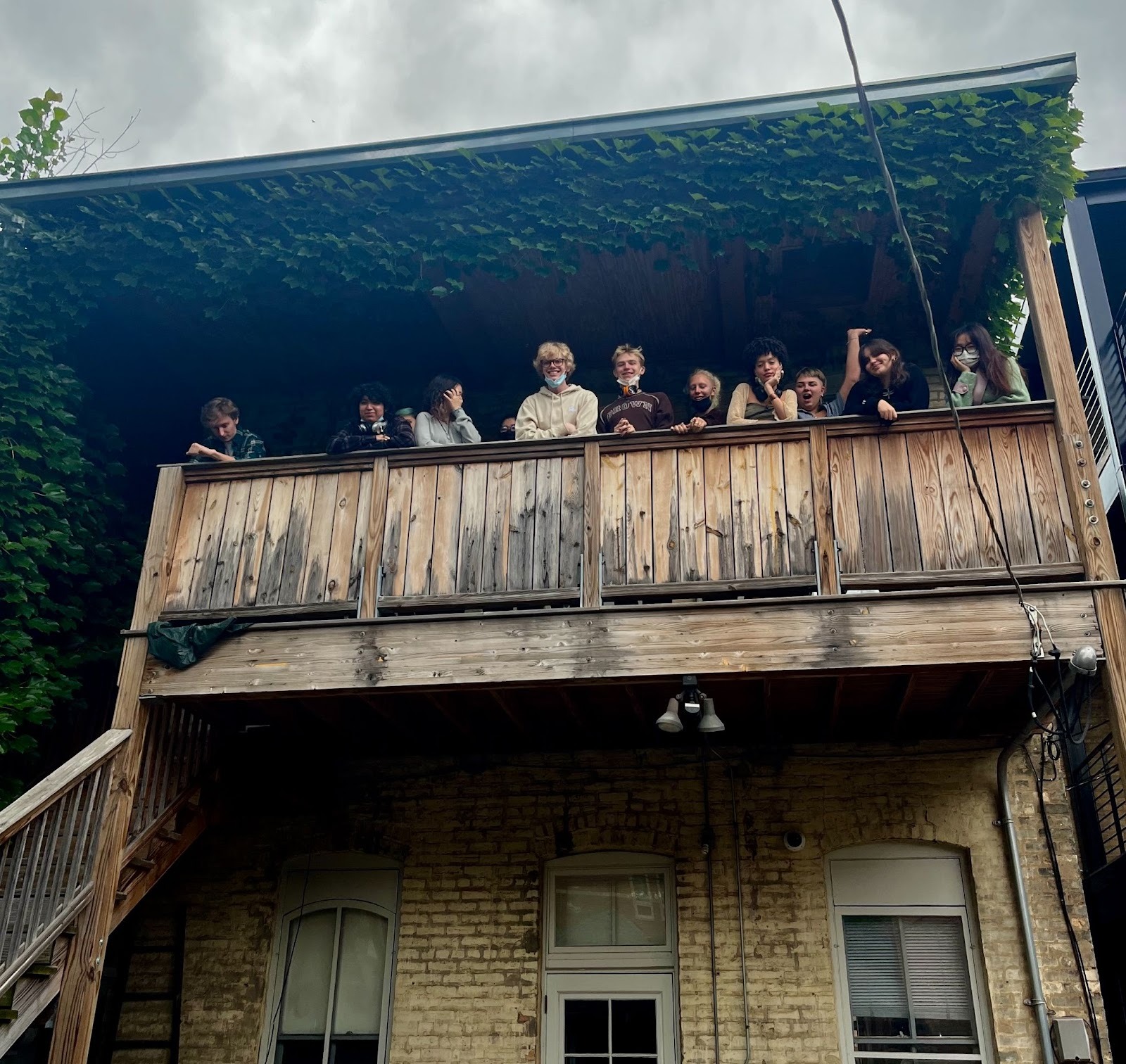 Image: Students from the School of the Art Institute of Chicago stand next to each other, forming a line across the railing of the wooden back porch platform which leads to Roger Brown's Study Collection in Chicago. The students smile as they pose for the camera. Green ivy lines the roof which covers the balcony.