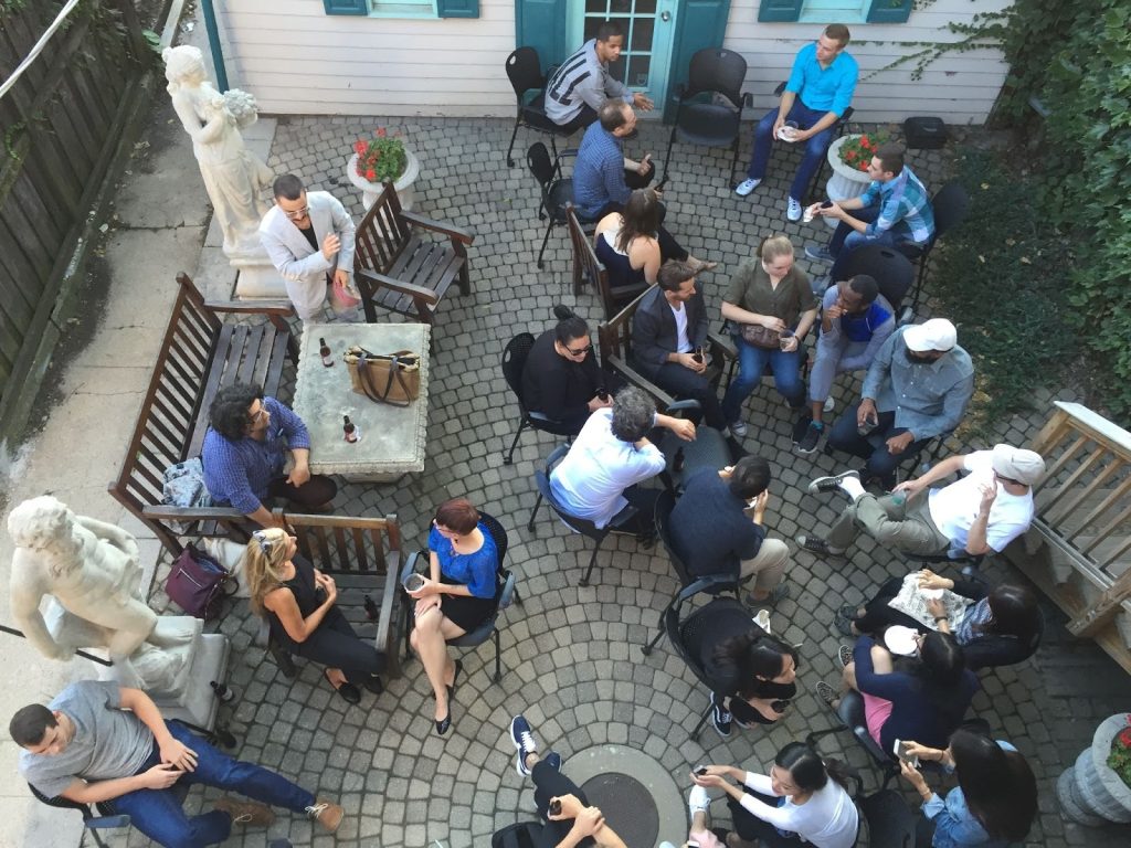 Birds eye view from the upper balcony of a backyard gathering at the Roger Brown Study Collection. The brick patio contains s dozen or so people are gathered on a sunny evening. The casually dressed people are sitting standing and small groups, conversing over beer and drinks.