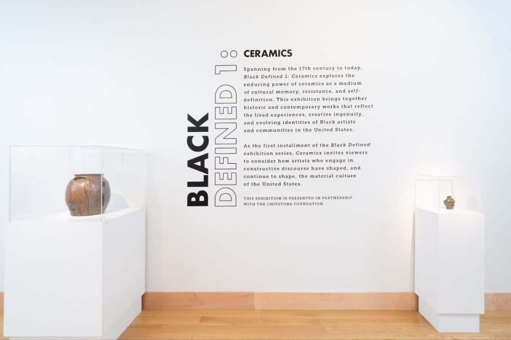 Installation View, “Black Defined 1: Ceramics,” 2025. Milwaukee’s Bronzeville Center of the Arts. Flanked on one side of the title wall is David Drake’s pot in a vatrine on a white plinth in a glass case, and on the other side is a face jug on a white plinth in a glass case. Photo: Daniel McCullough.