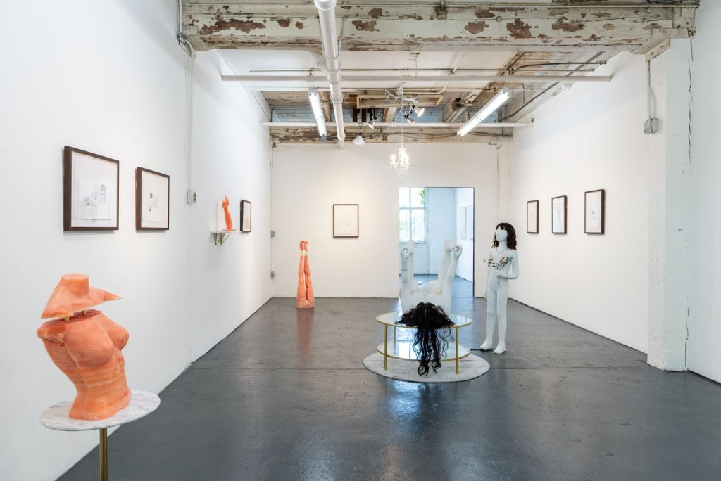 Installation view, Iris Bernblum: VARIOUS PLEASURES, 2023. Throughout the gallery space are figures of bodies. A terracotta-colored wax torso with a floating collarbone and shoulders is closest to the viewer. In the middle of the space, a white figure with black hair stands holding flowers while a second white figure with black hair lies on their back holding their feet up in the air. At the very back is a wax figure of legs upside down so the feet are pointed towards the ceiling. Around the gallery walls is framed artwork. Photo by Ry Thiel. Courtesy of Goldfinch.