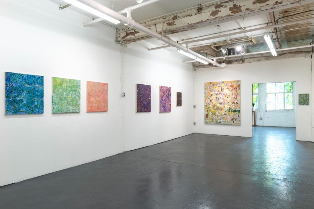Image: Installation view, Mari Eastman: Studio Simulacrum, 2017. Hung on the white gallery wall are six textured canvases. One blue, one green, one coral, one dark purple , one light purple, and one brown. They lead towards a larger, multicolored painting hung on the farthest wall. Courtesy of Goldfinch.