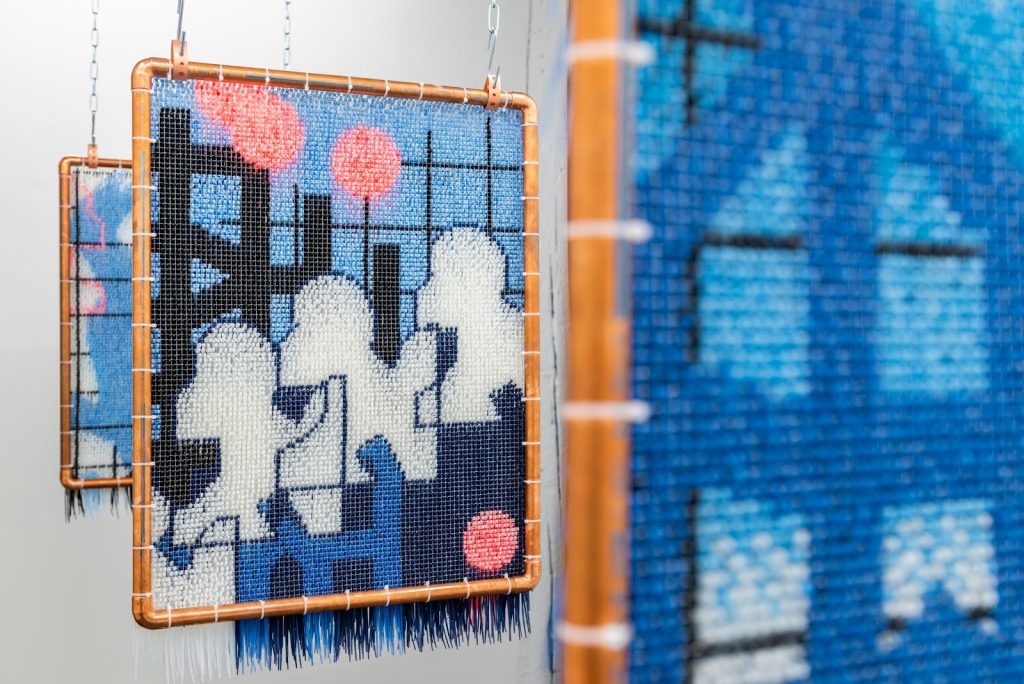 Installation view, Sarah Leuchtner: Sequential Escapes, 2023. Three works made of woven, colored zip-ties in geometric shapes of blue, white, and coral, are hung from the ceiling with metal chain. Each piece is secured inside a copper frame and the zip-tie ends hang from the bottoms like fringe. Photo by Ry Thiel. Courtesy of Goldfinch.