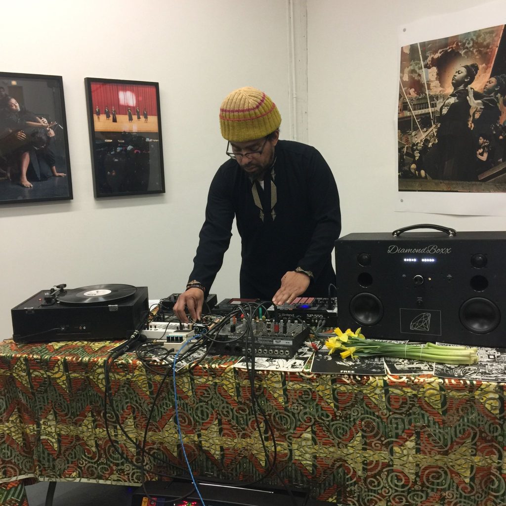 Damon Locks during the opening reception for Then X Enters, 2021. Locks is poised above a table filled with audio equipment and is mixing the sound that accompanied a performance by dancers Move Me Soul. (Dancers are not pictured in this shot, but images of these dancers are portrayed in the framed photographs behind Locks on the left.). Photo by Claudine Isé. Courtesy of Goldfinch.