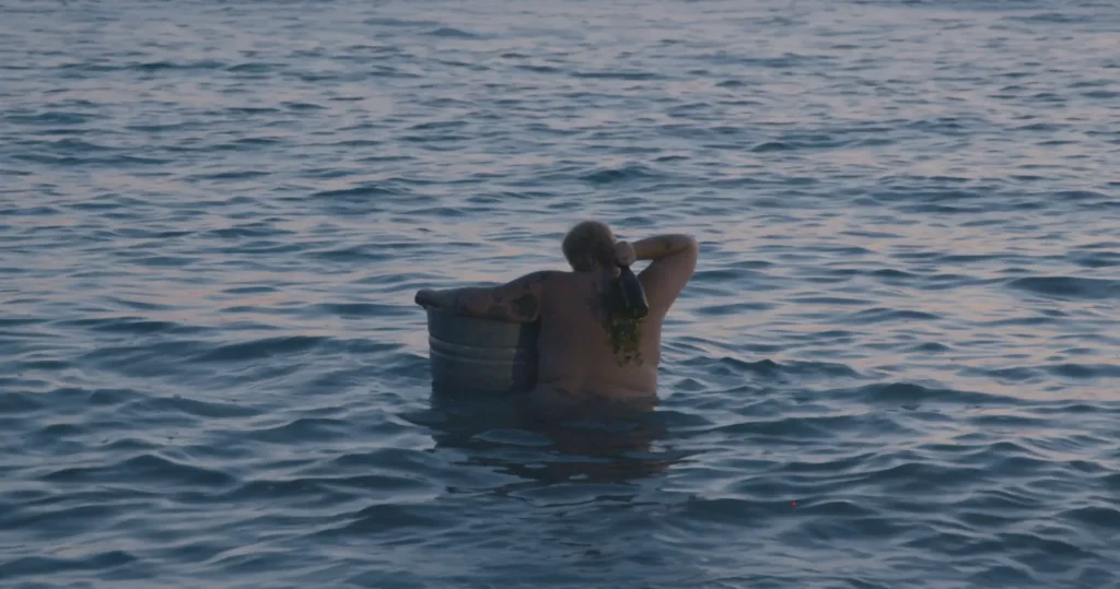 Detail shot of Maiden Voyage (2025). A naked person is waist deep in water holding a tin bucket underneath their left arm while smashing a champagne bottle over their right shoulder. Photo courtesy of the artist.