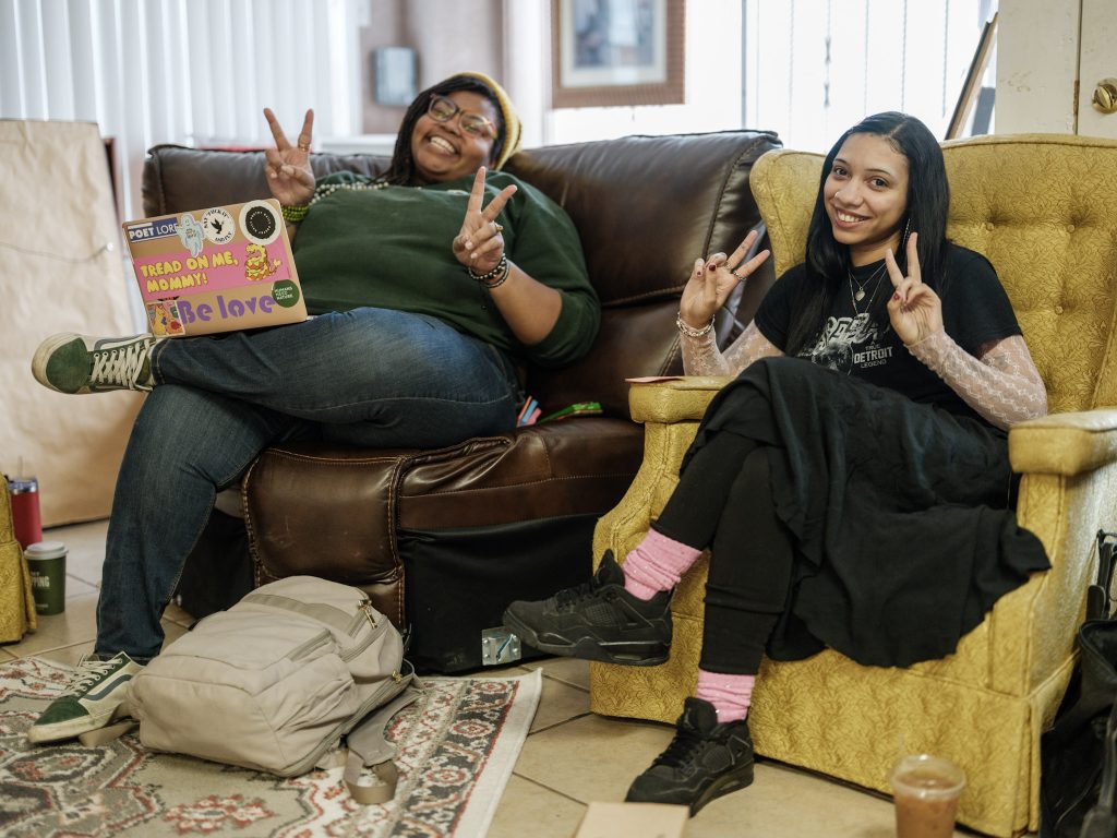 Image: Two folks sit next to each other at The KC Defender's Fall retreat at Willa’s Books & Vinyl. They both smile while holding up peace signs: on the left is Melissa Ferrer-Civil (Director of B-REAL Academy), and on the right is Nina Kerrs (Willa's Campaign Lead + Defender HQ Site Manager). Photo made by Vaughan Harrison.