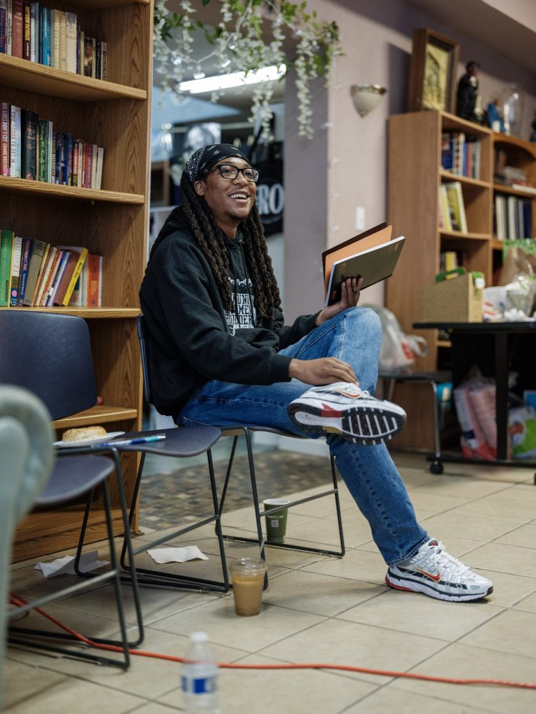 Image: Ryan Sorrell sits in a chair at The KC Defender's Fall retreat, which took place at Willa’s Books & Vinyl. He is smiling and holding an notebook. Photo made by Vaughan Harrison.