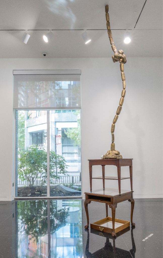 Installation view: Temitayo Ogunbiyi, You will have revelations along felled branches and longer roots/routes, 2019. A tall, golden sculpture made of discrete oblong pieces stretches from the top of wooden side tables to the ceiling of the exhibition space. Courtesy The Arts Club of Chicago. Photograph by Michael Tropea.