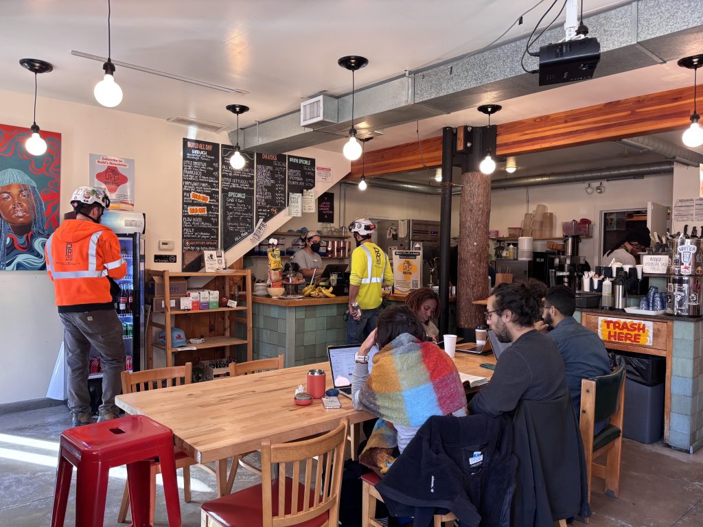 Image: A view of the front of Build Coffee & Books in Chicago. Folks sit around a table inside Build drinking coffee. Behind them, customers order from the teller behind the counter. Image courtesy of Andrea Faye Hart.