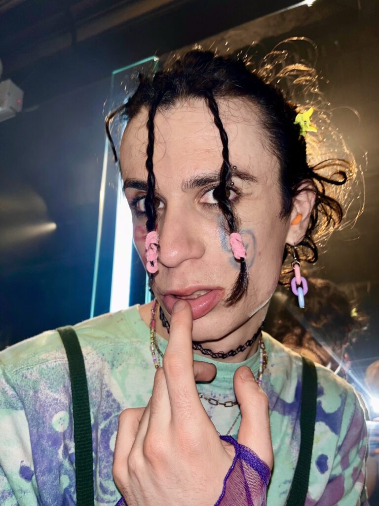 Image: Noa Fields dressed to dance at Smartbar club in Chicago. She wears her hair in mini braids with colorful hair ties and clips, colorful earrings, choker necklaces, a tie dye shirt, make up, and hand drawn face tattoos on each cheek. Image by Eva Geczy. 