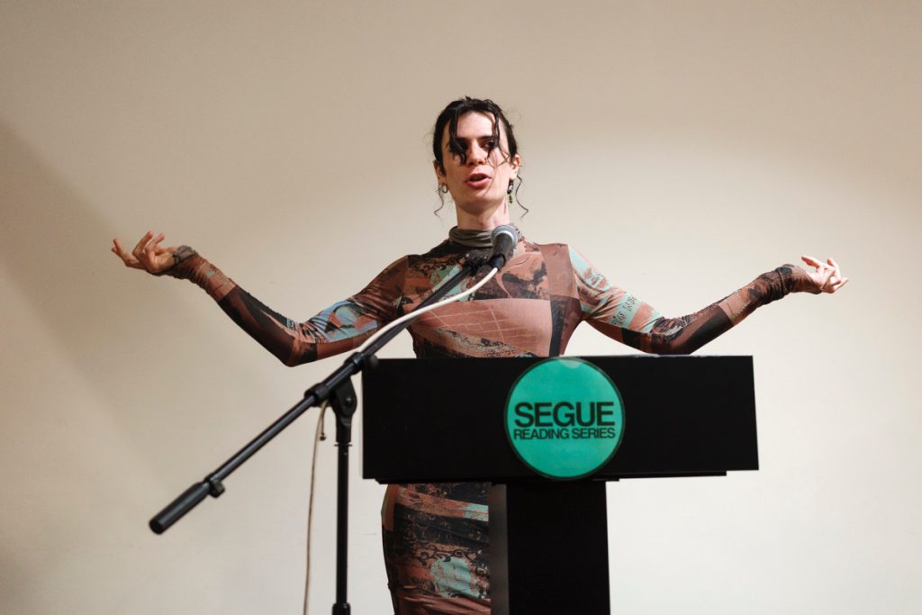 Image: Noa Fields wearing a colorful long-sleeved mesh dress stands by the microphone and pulpit during her reading at Segue Reading Series in New York City. Image by Ang Zheng.