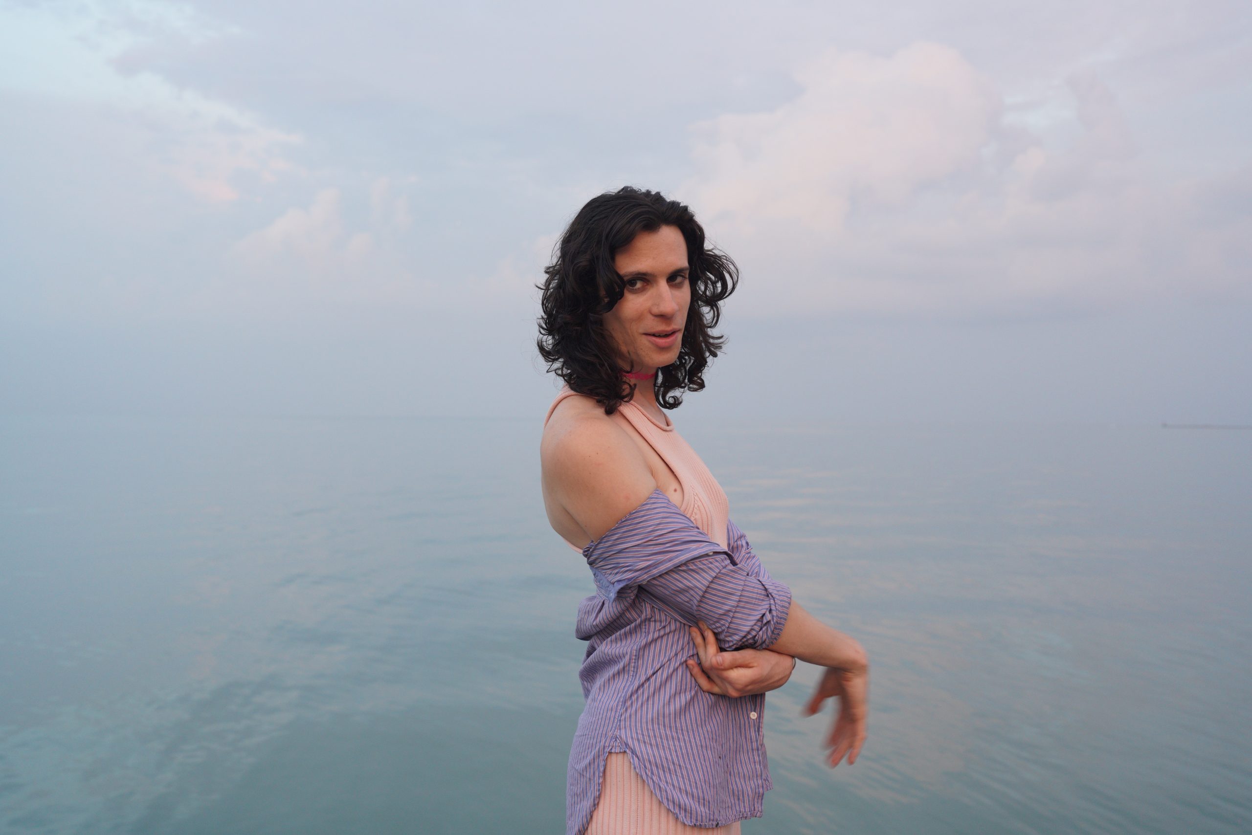 Image: Noa Fields, wearing a light pink swimsuit and purple button-down poses with her right side profile towards the camera next to Lake Michigan on a hazy afternoon during summer 2023. Image by Armando Lozano (IG: Hole Boss).