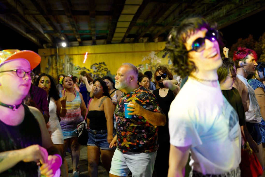 Image: Noa Fields, wearing dark sunglasses and a white graphic tee dances at Tropitecha pop up nightlife club. The dance floor is full of others enjoying the music. Image by Armando Lozano (IG: Hole Boss).