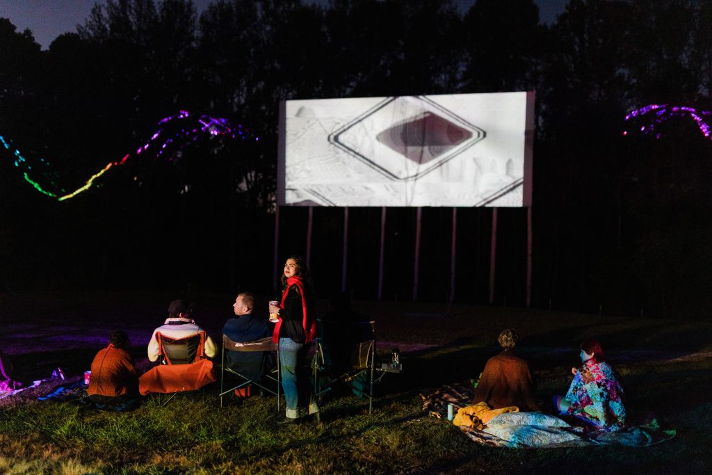 Image: A nighttime photograph that centers an attendee of "Signal Overload" looking back at the camera while standing in front of the screen while other attendees are seated during the show. Photo by Garrett Ann Walters.