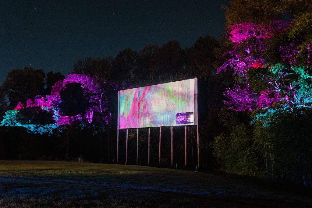 Image: A nighttime photograph captures colorful lights projected on both the screen and surrounding trees at Star Lite Drive-In Theatre. Photo by Garrett Ann Walters.
