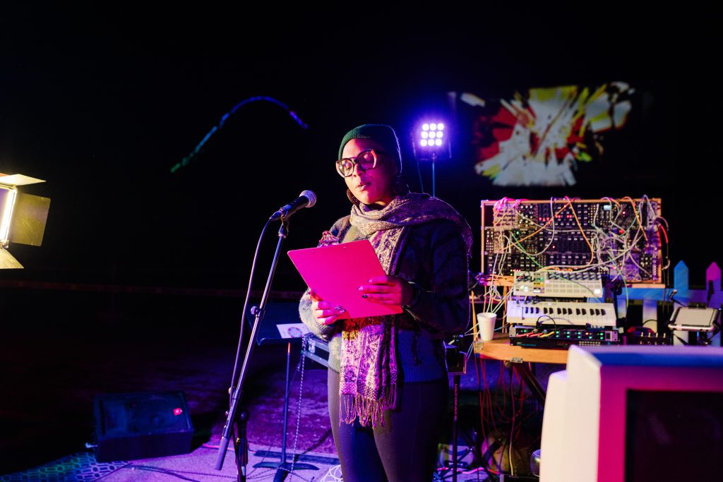 Image: Nighttime photograph of a performer at "Signal Overload" holding a clipboard with the screen and sound equipment behind them. Photo by Garrett Ann Walters.
