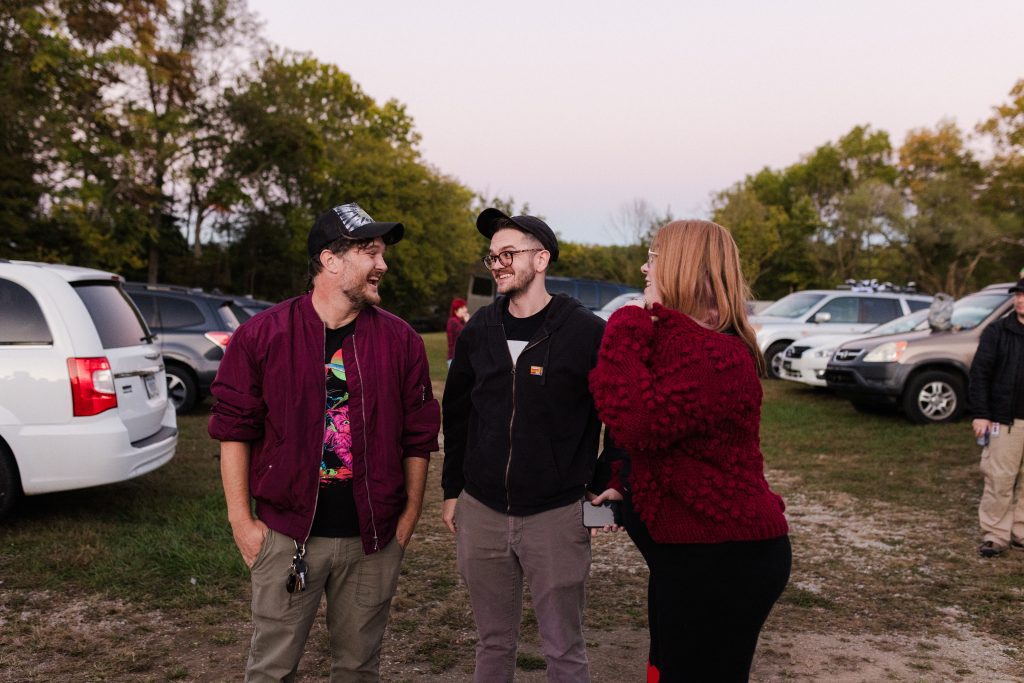 Image: Daytime photograph of three attendees of "Signal Overload" in mid conversation with each other while standing in between rows of parked cars at Star Lite Drive-in Theatre.  Photo by Garrett Ann Walters.