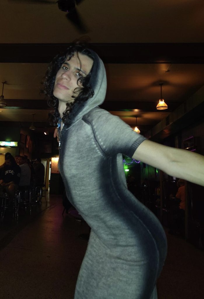 Image: Noa Fields wearing knit gray and black hooded body con dress poses with her arms stretched at her rebirthday at Skylark bar. Image by Mahnu.