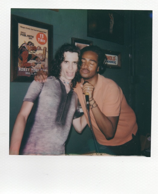 Image: Noa Fields who wears a knit gray and black body con dress poses next to her friend Jared Brown who wears a pink shirt, headphones, and is holding a microphone. Image by Jared Brown.