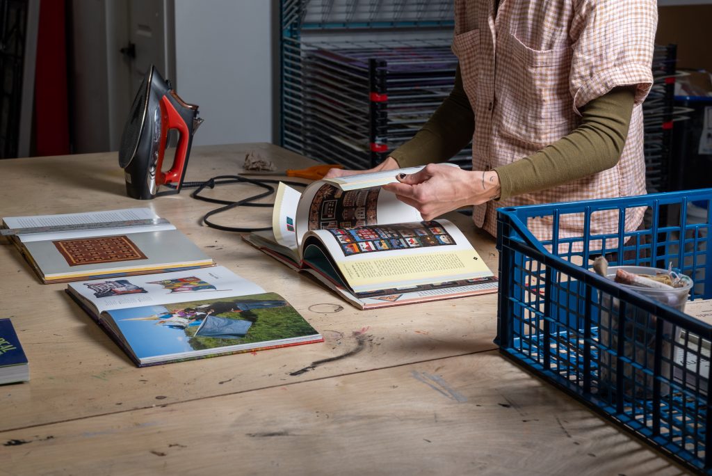 A close-up image of several books on a tabletop alongside a blue crate of supplies and an electric iron. A woman flips through one of the books. Image by Tonal Simmons.
