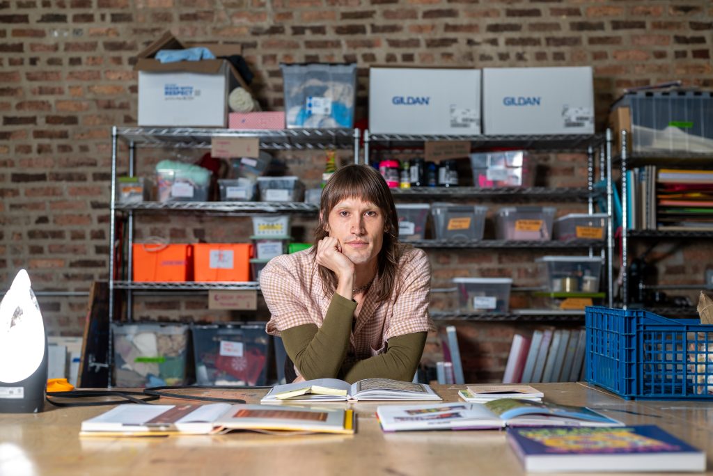 A woman poses in front of shelves stocked with supplies and with a number of books displayed on the table in front of her. Image by Tonal Simmons.