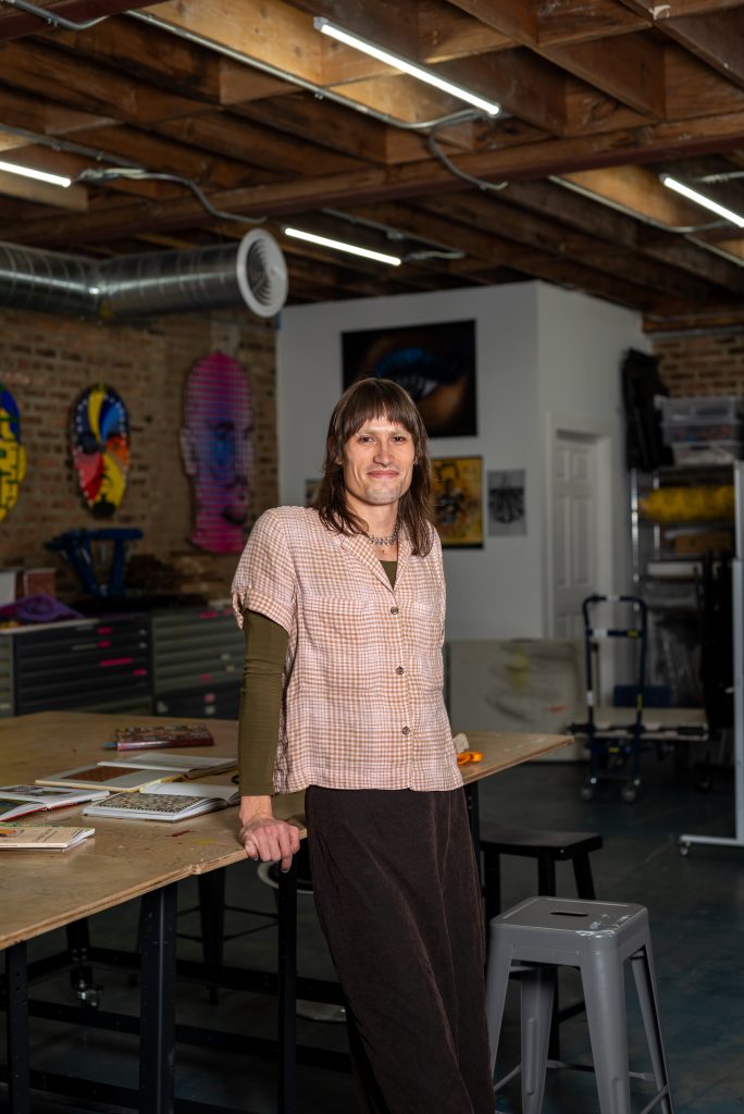 A portrait of Eve Emrich. A woman with mid-length brown hair wearing a button-up over a long-sleeve t-shirt leans against a table with a number of books on it. Artwork and tool cabinets are visible behind her. Image by Tonal Simmons.