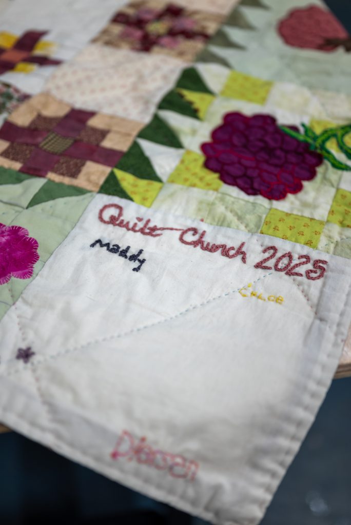 A detail image of a white, corner quilt square with hand-embroidered text that reads "Quilt Church 2025," in addition to several names. Other quilt squares in view feature designs created with fabric in many shades of purple, green, and peach. Image by Tonal Simmons.