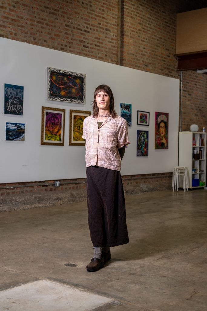 A portrait of Eve Emrich. A woman wearing a long brown skirt and button-up stands in the center of a room with a number of paintings mounted on the wall behind her. Image by Tonal Simmons.