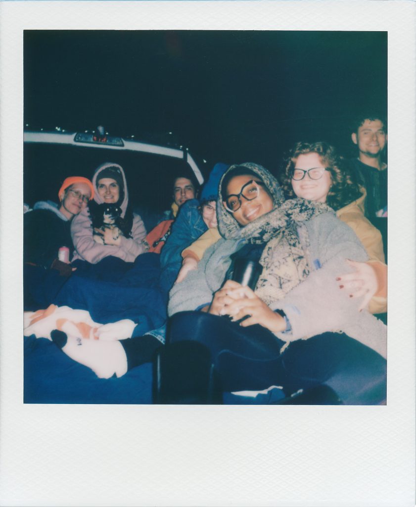 Image: Polaroid capture of seven attendees enjoying "Signal Overload" in the bed of a white pickup truck. Photo by Garrett Ann Walters.