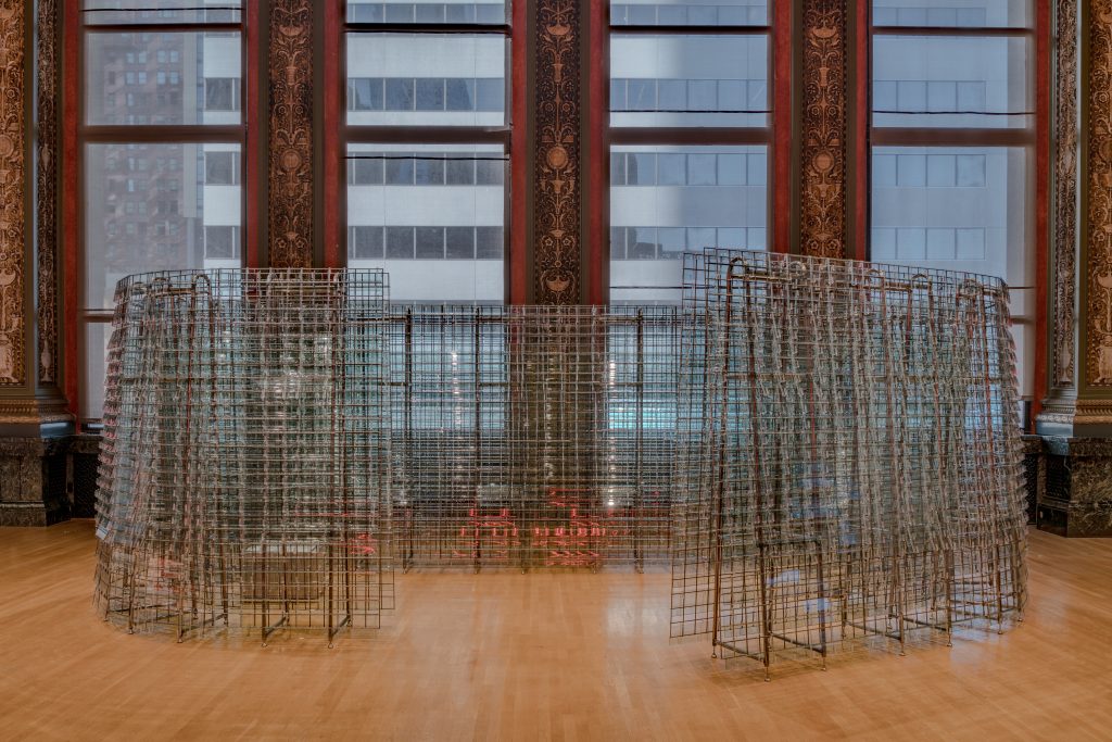 Installation view of Our Second Skin, the Skin of the City, RADDAR (São Paulo, Brazil) shown on the 4th floor of the Chicago Architectural Center at Chicago Architectural Biennial. The piece consists of 2,500 glass pieces, shaped like fish scales,  that altogether form a curved, translucent structure. Behind the structure Chicago Cultural Center's large windows are in view. Image courtesy of Chicago Architectural Biennial.