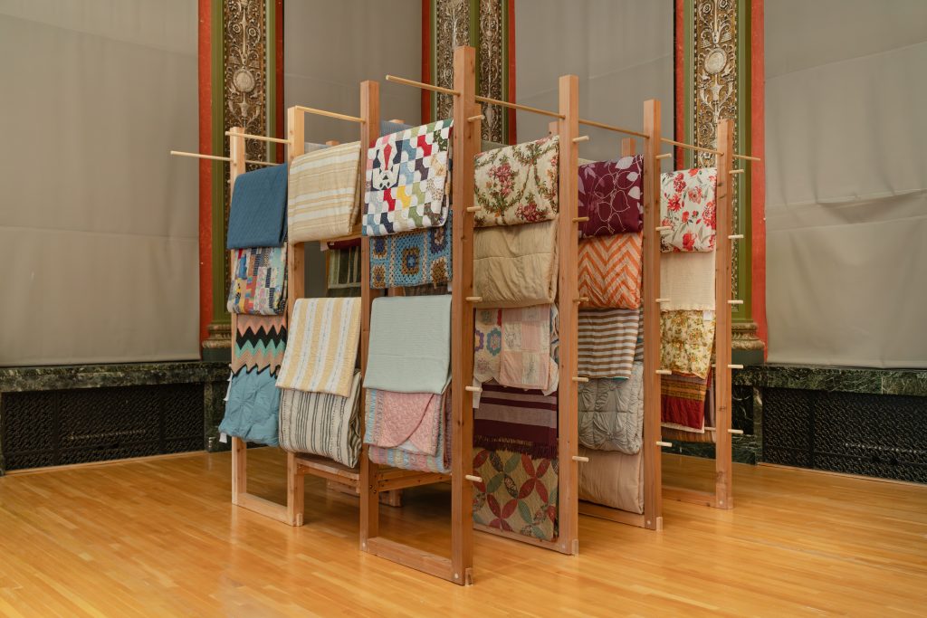 Installation view of The Linen Closet by Jason Campbell / ellProjects (Chicago, United States)  in which colored quilts an linen line a wooden structure, where they each fold over a thin wood beam. Image courtesy of Chicago Architectural Biennial.