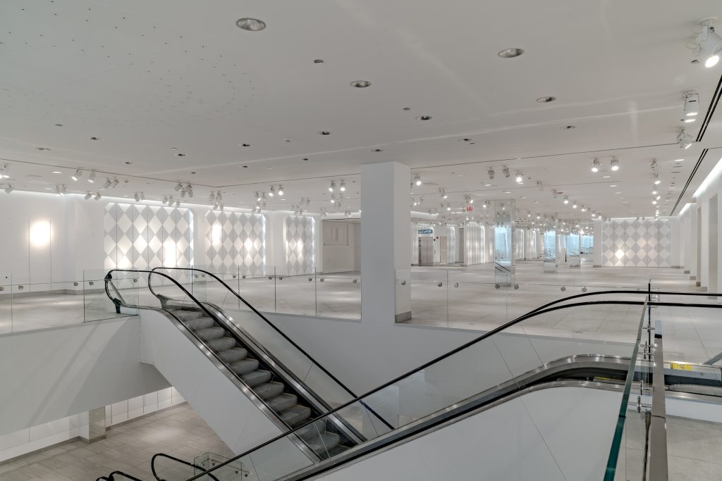 Indoor view of 840 N. Michigan as displayed in the Sixth Architectural Biennial. The view of the building is empty and all white except for gray patterning on the walls. Two escalators are shown running in the foreground of the image which is brightly lit by the space's built in light fixtures. Image courtesy of Chicago Architectural Biennial.