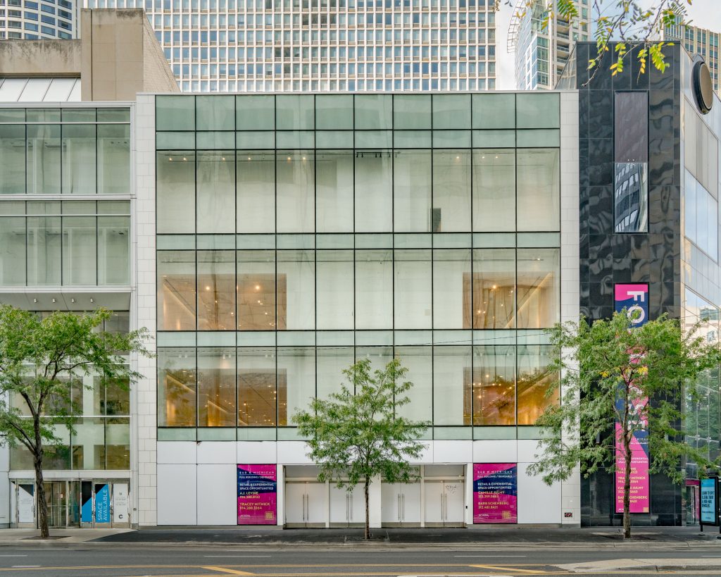 Exterior view of 840 N Michigan as displayed in the Sixth Architectural Biennial. On a sunny spring day and a clear street, we see a front view of a square shaped 4-story building with opaque white doors on its first floor and glass windows on each floor above. Yellow light is shown on the 2nd and 3rd floors from inside the building. Image courtesy of Chicago Architectural Biennial.