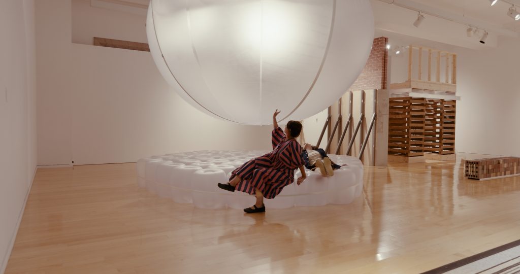Installation view of Surfaces in Flux Objects of Common Interest/LOT office for architecture (Athens, Greece and New York, United States) showing an clear inflatable mattress with clear large inflatable orb above it. A gallery-goer sits on a mattress wearing a striped red and black dress while touching the orb. Another gallery goer lays on the mattress behind the other. The piece "Two Foundations" by 11 x 17 (Denver, United States and Toronto, Canada) can be seen in the behind the inflatables. Image courtesy of Chicago Architectural Biennial.