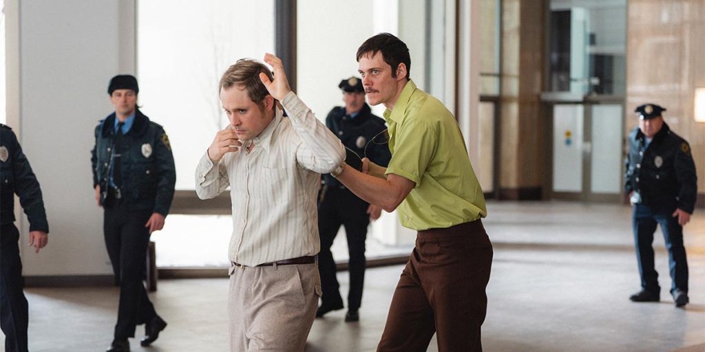 A frenzied man wearing a green shirt and brown trousers holds another at gunpoint inside the lobby of a building. Four uniformed police officers surround them prepared to draw their own guns.
