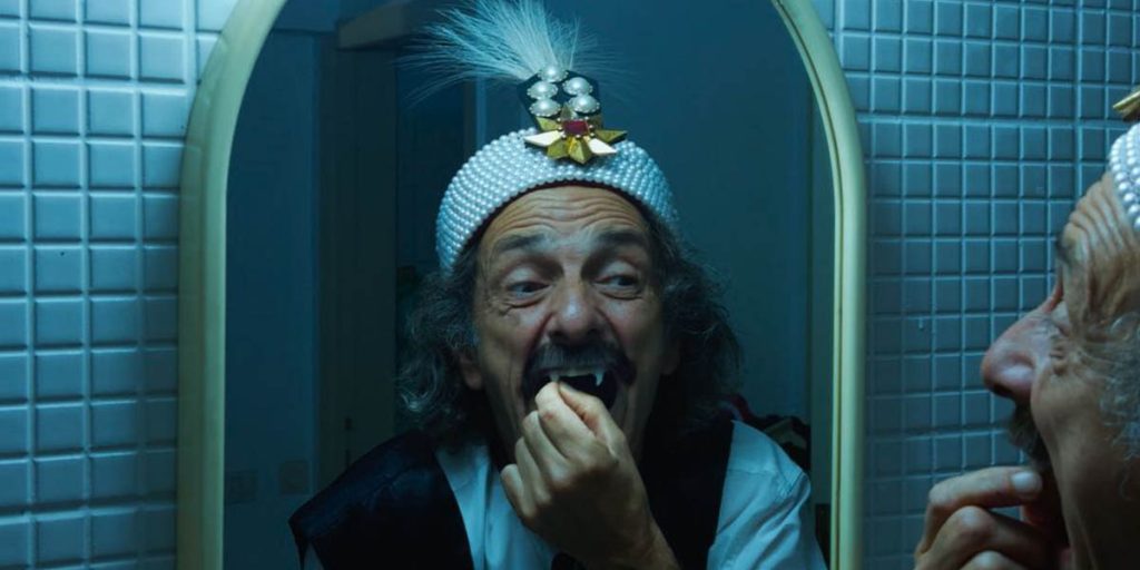 An aging man with long gray hair stands before a mirror wearing a pearled hat bedazzled with jewels and a feather. He is gripping his fangs with his thumb and index finger.
