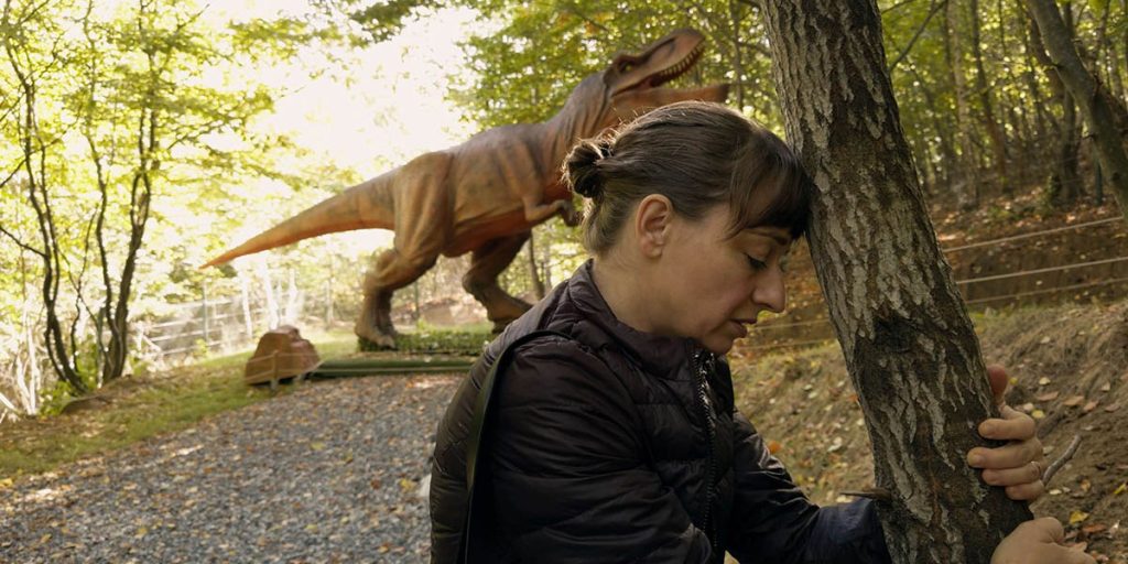 A woman grips the trunk of a tree and presses her forehead against it. She is wearing a light jacket and her hair is pulled into a bun. There is a T. rex statue behind her at the end of a gravel road surrounded by forest.
