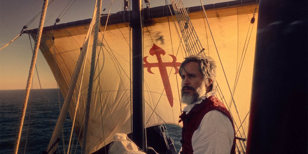 A man with gray hair and a beard looks out to the sea. A sail bearing the Spanish Order of Santiago cross is visible in the behind him.