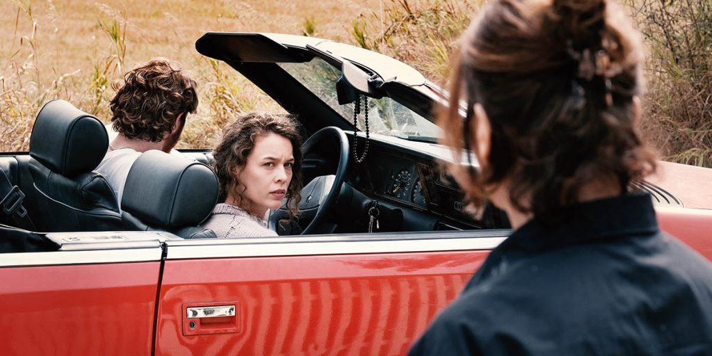 A couple are riding inside a red convertible. The man is facing away from the camera while the woman looks suspiciously at a woman in the foreground of the image.