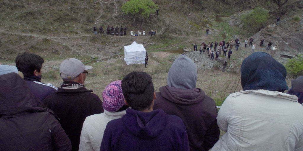 A crowd of people, including several who appear to be police officers or government officials are gathered in a valley near a white tent. A crowd of people watch from a hilltop with their backs to the camera.