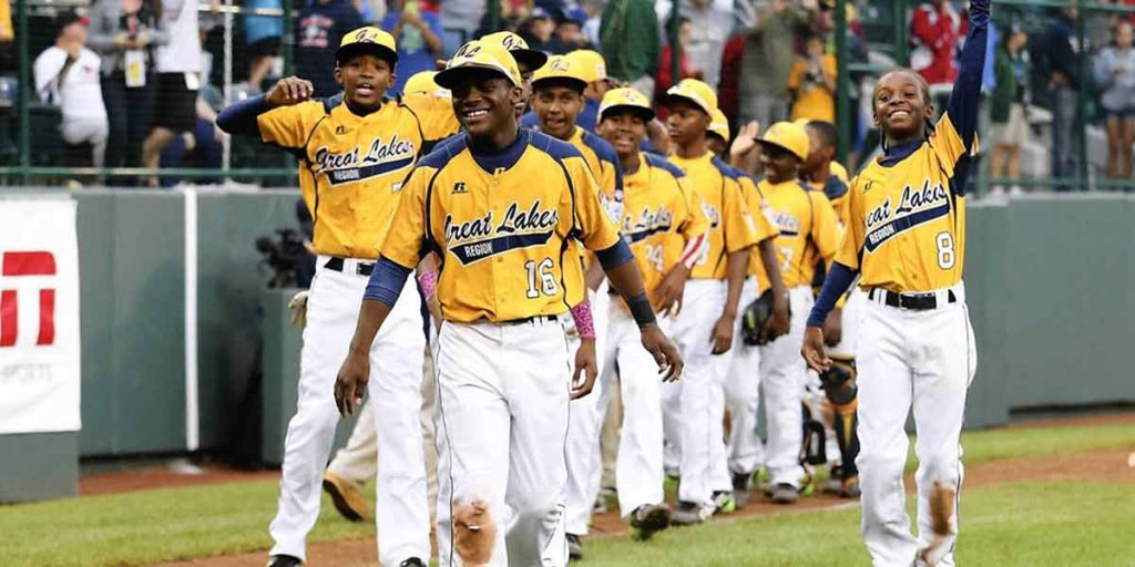 A group of baseball players march onto a field wearing yellow and navy jerseys with the words "Great Lakes Region" embroidered onto them. Two of the boys have dirt stains on the shins of their white pants. Baseball goers are visible in the grandstand behind the boys.