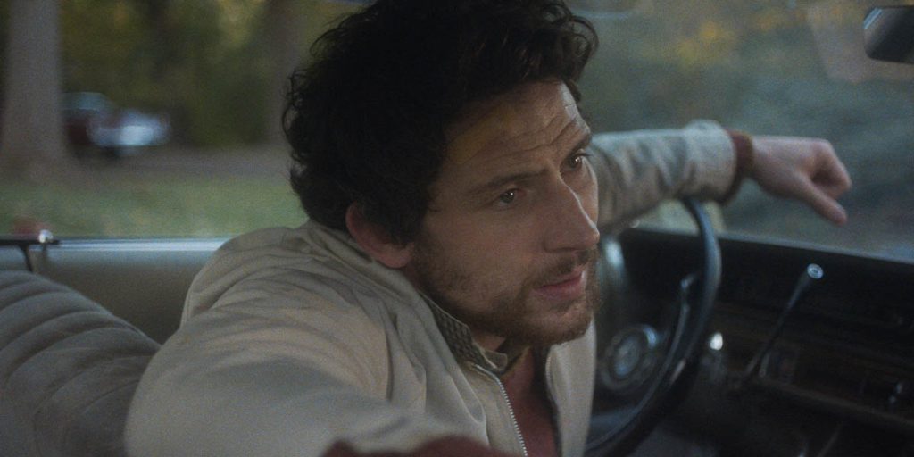 A closeup of a man inside his car. His forehead is wrinkled with concern as he looks out the side window with his arms resting on the steering wheel and seat.