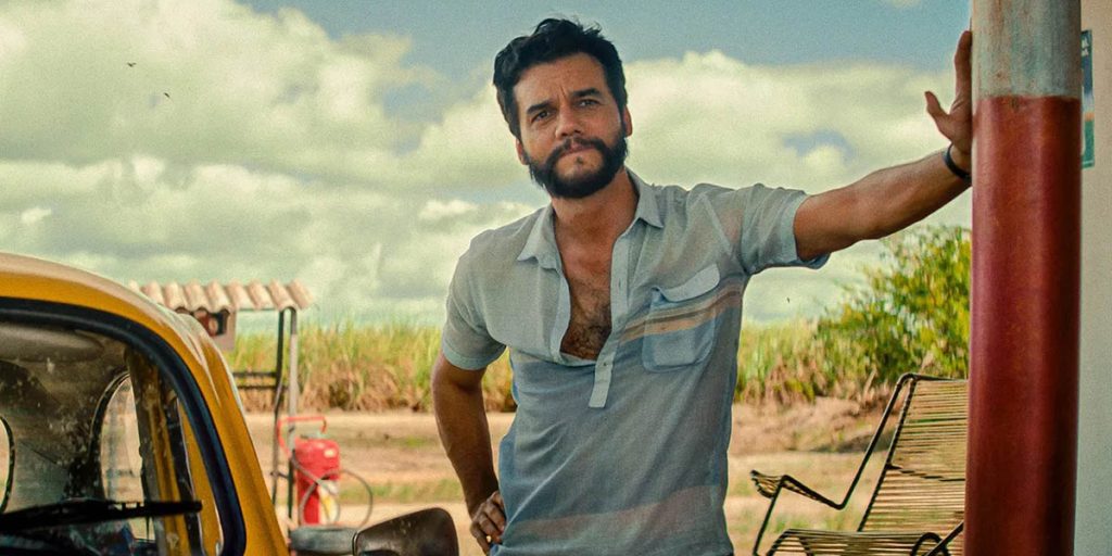 A man in an unbuttoned blue polo stands beside his car and looks concerned while resting his hand on his hip and leaning against a pillar. A field of reeds are visible in the background.