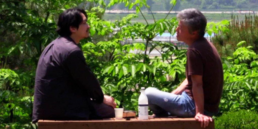 Two men are seated on a bench facing each other. A lake is visible in the background. A wall of plants separates the men from the waterfront. There is a bottle and a cup between them.