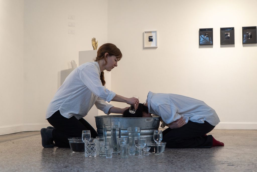 A red-haired woman with a white button-up shirt and rolled sleeves gently pours water from a glass onto another person’s short black hair. They both face each other and kneel over a metal bucket in the middle of a gallery space. There is a group of variously-shaped glasses full of water in front of the bucket.  